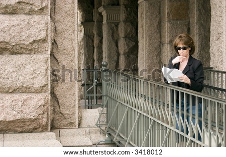 A woman lawyer standing on a ramp leading to a large building reading something on a few sheet of paper she is holding in her hand.
