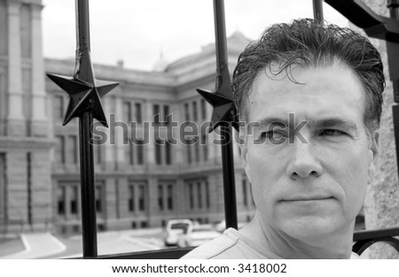 Black and white photo of a man standing in front of a gate with a crafty look  on his face.