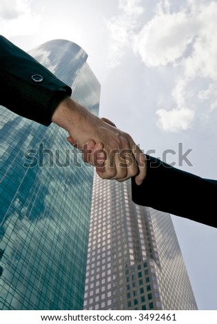 A handshake between a man and woman with tall beautiful glass towers of commerce in the background.