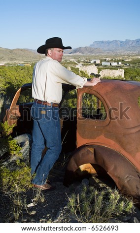 A man in western attire leaning on an old, long abandoned vehicle surrounded by rugged terrain.
