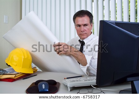 A man sitting at a desk with a hard hat and computer holding building plans or blueprints in his hands.