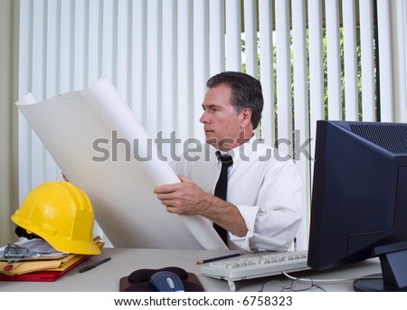 A man sitting at a desk with a computer and a hard hat holding building plans or blueprints.
