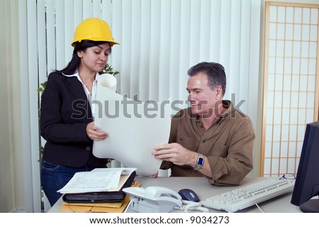A young woman in a hardhat and a man looking over what appears to be construction plans.