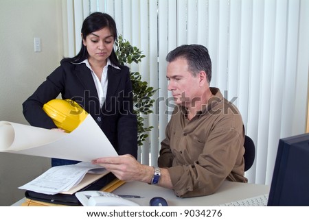 A woman holding a hardhat looking at what appears to be a set of plans held by a man.