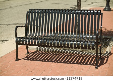 The harsh sun casting shadows from an empty, slotted, black, metal bench.