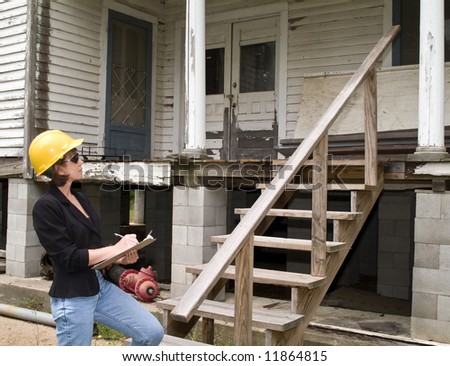 A woman in a hard hat, holding a clipboard, standing in front of an old house.