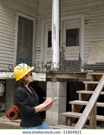 A woman standing in front of an old rundown house, wearing a hard hat and writing on a clipboard.
