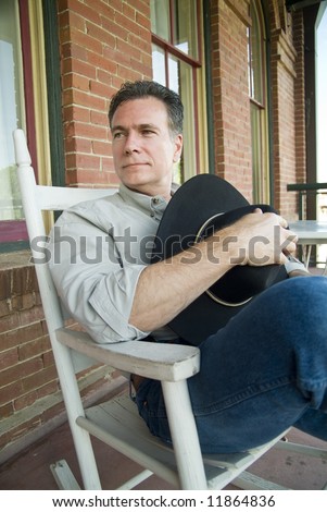 A man with a friendly smile, sitting on a porch in a rocking chair, holding a cowboy hat.