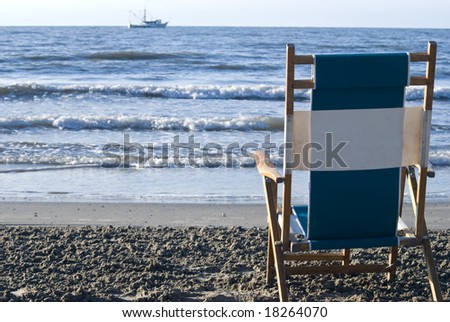 View of the surf from behind a single beach chair with a fishing boat in the distance.
