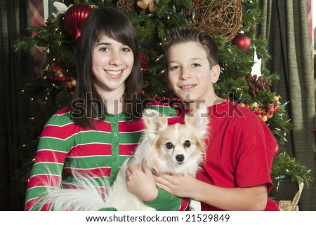 Two smiling children in front of a Christmas tree holding their little pet dog.