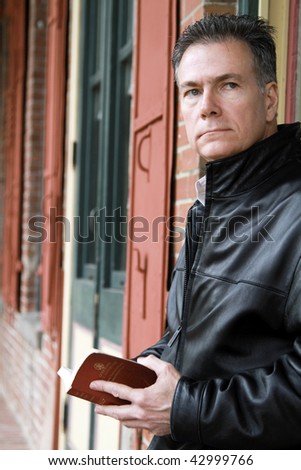 A pensive man holding a New Testament bible, standing by a building with colorful windowpanes and shutters.