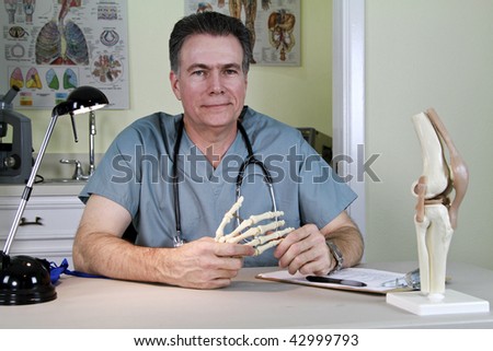 An smiling orthopedic doctor holding a skeletal model of the human hand, with a model of a human knee on his desk.