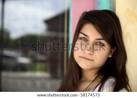 A beautiful brunette girl with piercing eyes gazes at the camera as she leans against a colorfully painted wall.