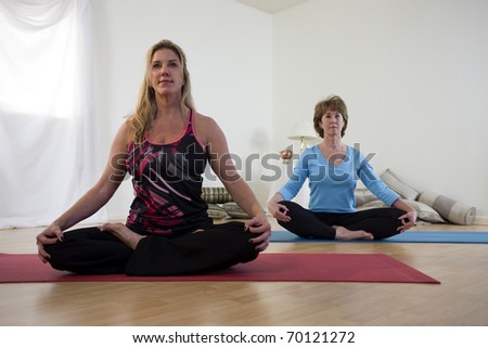 A yoga instructor and her student seated crossed leg on their mats in a small studio prepare for yoga practice.