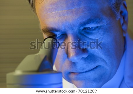 A blue light covers the face of a man who may be a scientist or doctor looking into a microscope. Blue gel used to create affect.