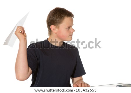 Teenage boy seated at a desk holding a paper plane above his shoulder taking aim at one of his classmates isolated on white