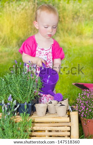 Cute two years old baby girl in a pretty pink dress standing outdoors in the garden watering the plants with a small purple plastic toy watering can