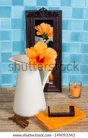Bathroom still life with a beautiful exotic orange hibiscus flower in a jug reflected in a small mirror leaning against turquoise blue tiles with a burning candle, towel and nail brush