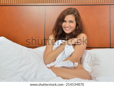 Beautiful brunette woman sitting on bed covering herself with a sheet and with smile