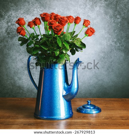 Red roses bouquet in glossy blue retro coffee pot on wooden surface behind grey wall background