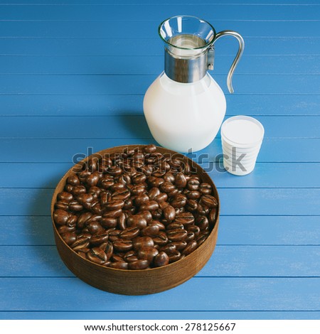 Vintage pitcher and glass full of organic bio milk and plate with roasted coffee beans on blue wooden surface. Picture of breakfast in cafe.