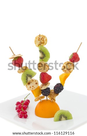 Mini pancake with cut fruits on skewers, fixed on half orange, decorated with red currant, on white plate