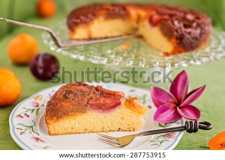 Slice of homemade upside-down plum cake on plate with fork and purple frangipani flower