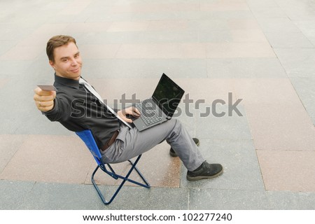 Image of the businessman sitting with the laptop and giving a card at the camera in the empty street. Focus is made on top of the background marbled tile in the street.