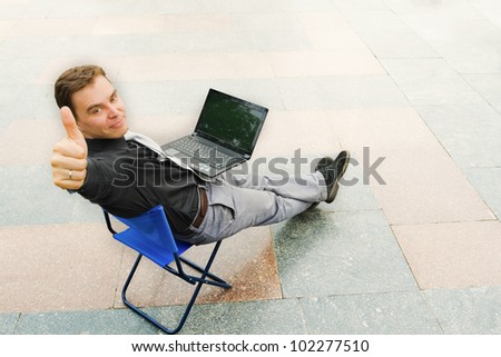 Image of the businessman sitting with the laptop and looking at the camera in the empty street. Focus is made on top of the background marbled tile in the street.