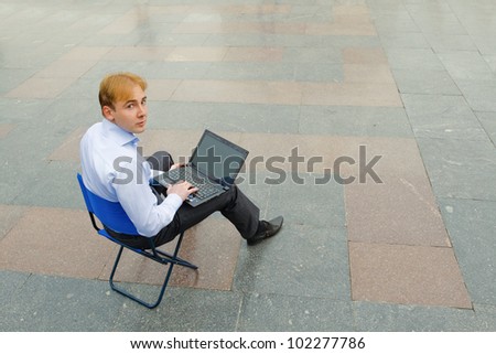 Image of the businessman sitting with the laptop and looking at the camera in the empty street. Focus is made on top of the background marbled tile in the street.