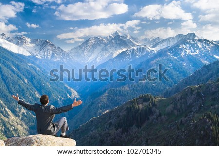 Image of a young businessman who sits on the top of the mountain on the background of the beautiful mountains, relaxing and pleased with the achieved successes.