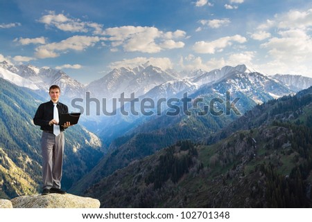 Image of a businessman standing on the top of the mountain, using a laptop and looking at the camera happy with his success. In the background a beautiful panorama of the valley with high peaks.