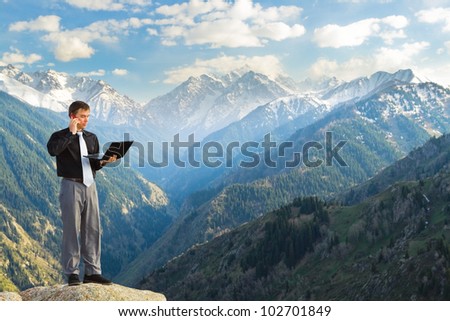 Image of a businessman standing on the top of the mountain, using a laptop, talks on the phone. In the background a beautiful panorama of the valley with high peaks.