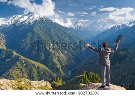 Image of a young businessman with laptop standing at the top of the mountain and looking into the distance to the beautiful mountains, is pleased with the achieved successes.