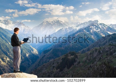 Image of a businessman standing on the top of the mountain, using a laptop. In the background a beautiful panorama of the valley with high peaks.
