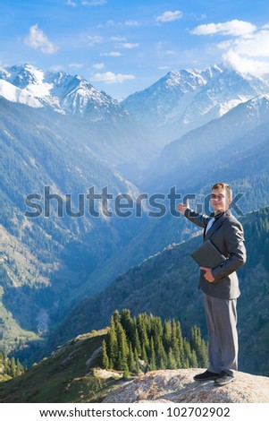 Image of a young businessman with laptop standing at the top of the mountain and looking at the camera and showing views of the beautiful mountains.