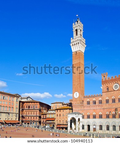 The vertical image of the main square of Siena, with a high tower and a clock and numerous number of tourists on a background of blue sky.