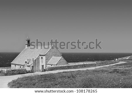 Black and white image of a stone church on the coast of Finistere, near the Cape of Ra. In the background are walking tourists, paying no attention to the strong north wind. Bretagne, France.