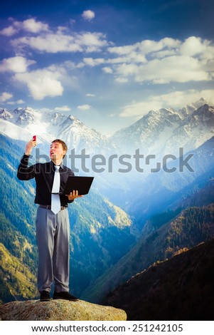 businessman standing on the top of the mountain, using a laptop and shouting into mobile phone