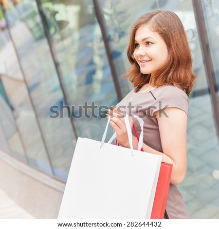 Happy shopping woman with a white bag for any text or label design, outdoor near the showcase of shopping center. Square orientation