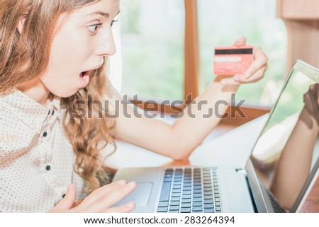 Beautiful surprised woman holding credit card, and shopping through laptop. She uses online payment by plastic card through the Internet Banking.