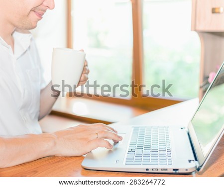close-up of young man dressed in a white shirt uses laptop at his workplace at home against window, he holds in his hand a cup of coffee