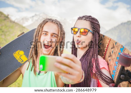 Happy people make selfie on mobile phone at mountain outdoor. They dressed at hippie style clothing, sunglasses, has long hair, holding skateboard and longboard