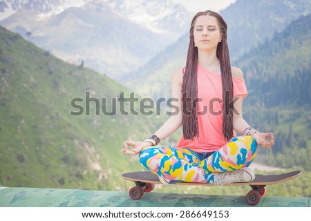 hippie fashion girl doing yoga, relaxing and sitting on skateboard at mountain. Concept of freedom and healthy lifestyle playing sports for yoga and skateboarding