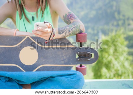 Closeup of hippie young and handsome man with skateboard outdoor at mountain. He using mobile phone and listening to music