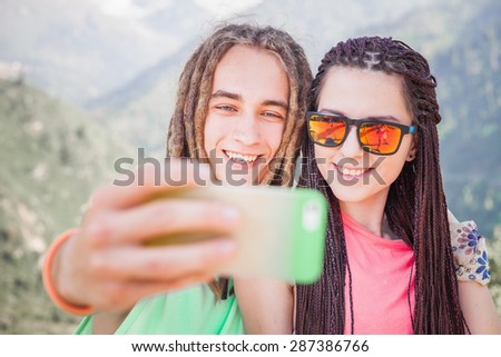 Happy and funny people make selfie or make pictures of themselves on mobile phone at mountain outdoor. They dressed at hipster style clothing, sunglasses, has long hair