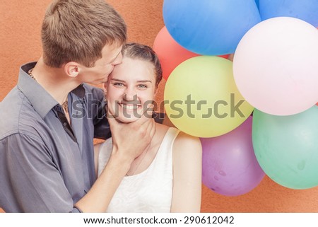 Young funny and unusual couple near the orange wall stand with balloons. Man holding the girl by the neck. Happy girl laughing and looking at the camera