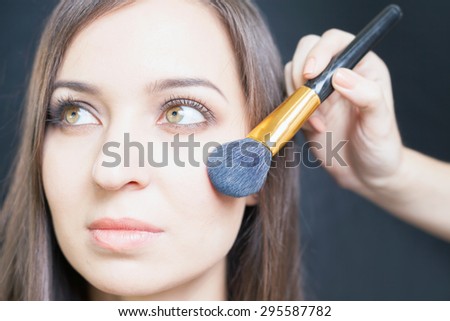 makeup artist doing make up using cosmetic brush for beautiful caucasian woman face at beauty salon with black background