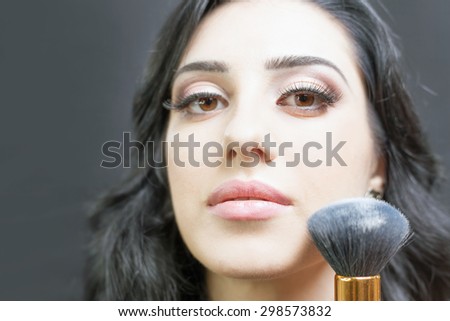 Beautiful arabian woman at beauty salon with a nice makeup. Make up specialist apply foundation cream on face, holding in hands a makeup brushe on a dark or black background.