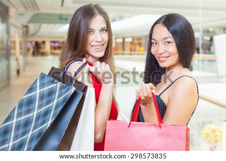 Two fashion girlfriends shopping at mall. They are looking at camera and smiling and hold handbag or bags at hands. Against the boutique